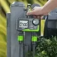 Close-up of a dual outlet garden hose timer mounted on a tap in a UK-style garden setting, with a digital screen visible and one hand adjusting the controls beside a grey rain barrel and greenery.